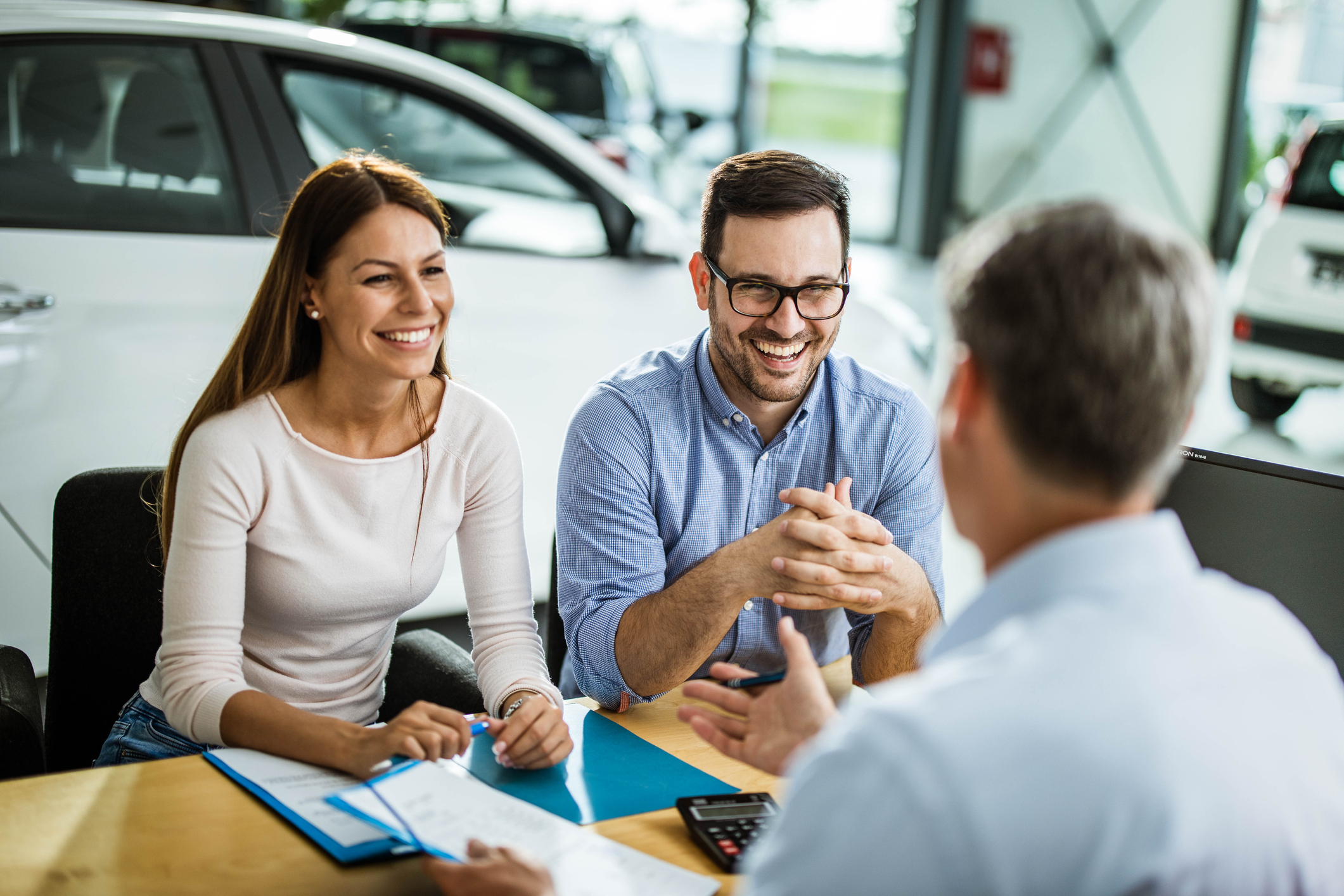 Young happy couple communicating with car salesperson on a meeting in showroom. Focus is on young man.