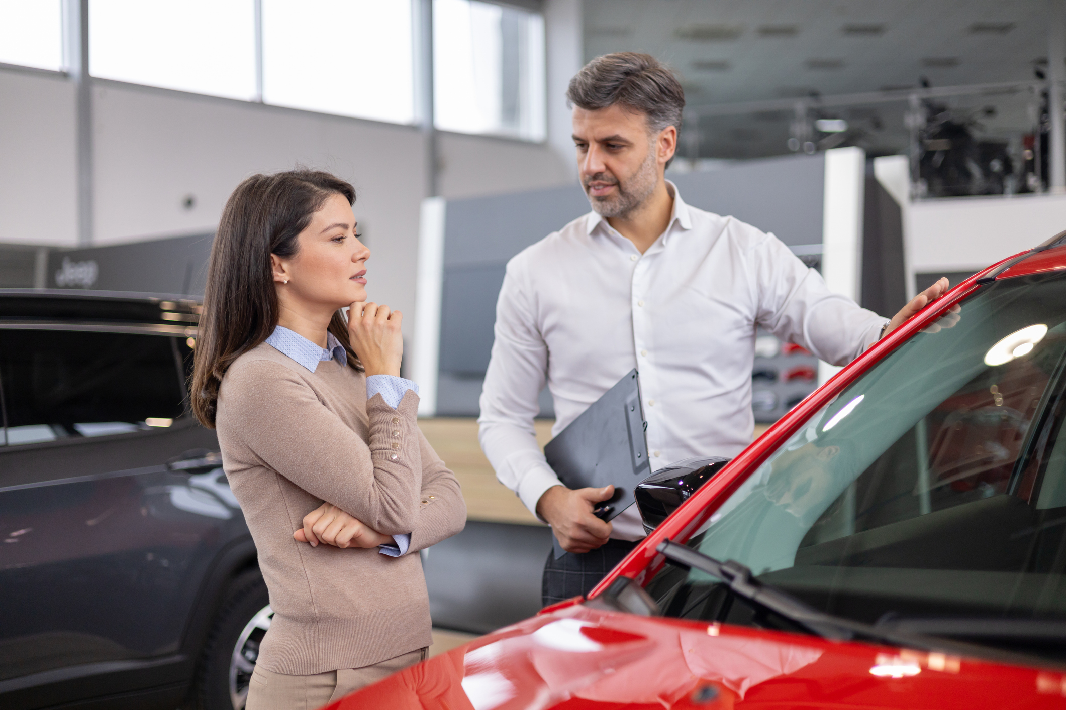 A man and a woman stand beside a red car as they purchase a new vehicle from a salesperson at a car dealership.