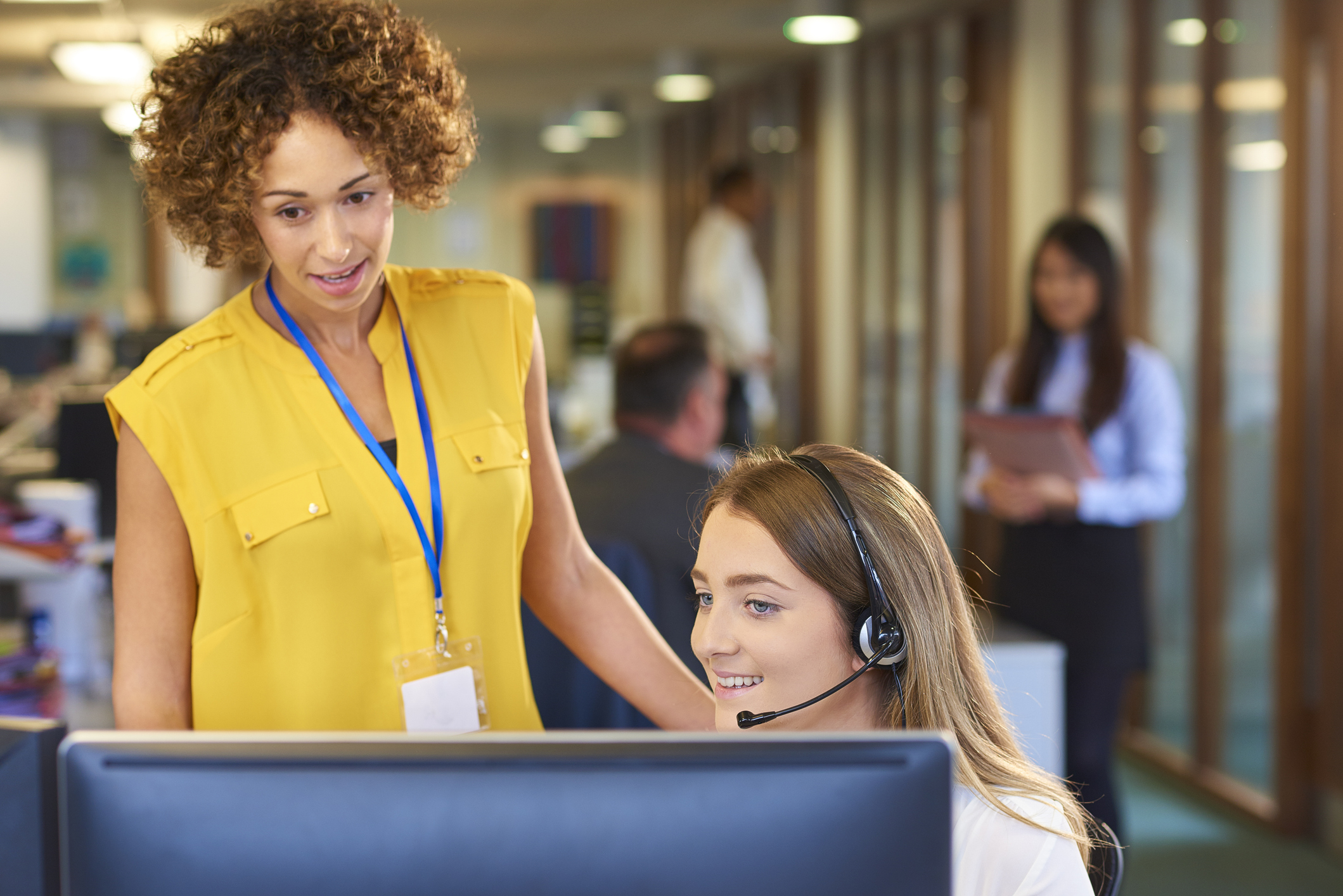 a young call centre representative greets a caller in a large open plan office as her supervisor watches over her shoulder and helps her through the call. Co-workers can be seen defocussed in the background .