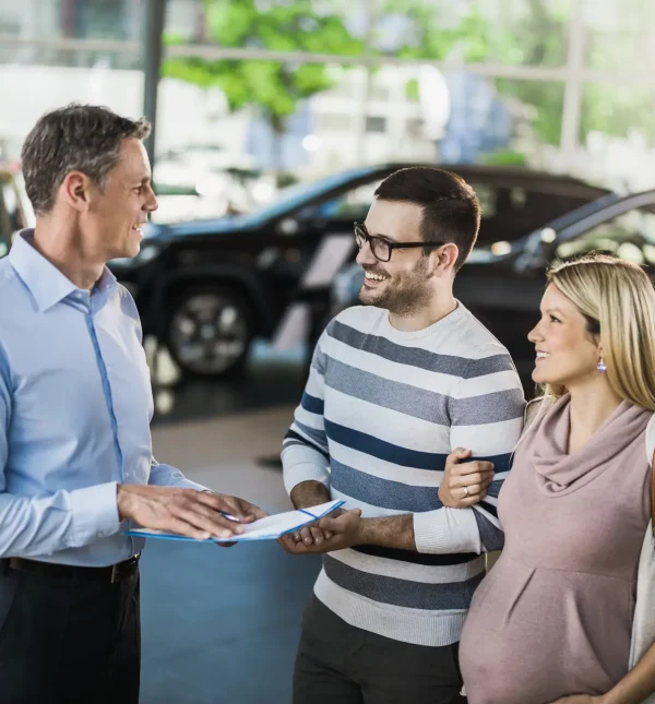 Happy pregnant couple talking to car salesperson in a showroom.