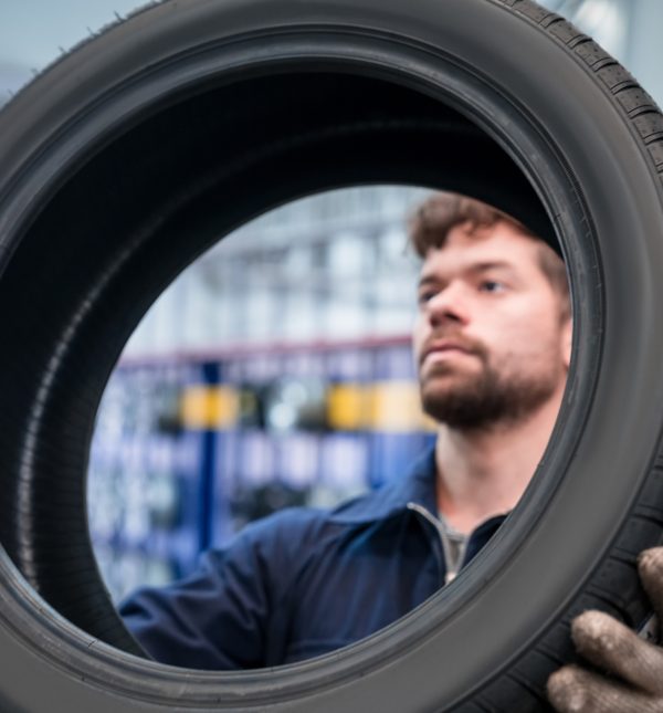 Young apprentice holding car wheel in the auto repair shop. Close-up of tire and face.