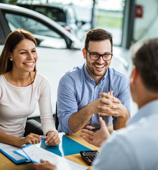 Young happy couple communicating with car salesperson on a meeting in showroom. Focus is on young man.