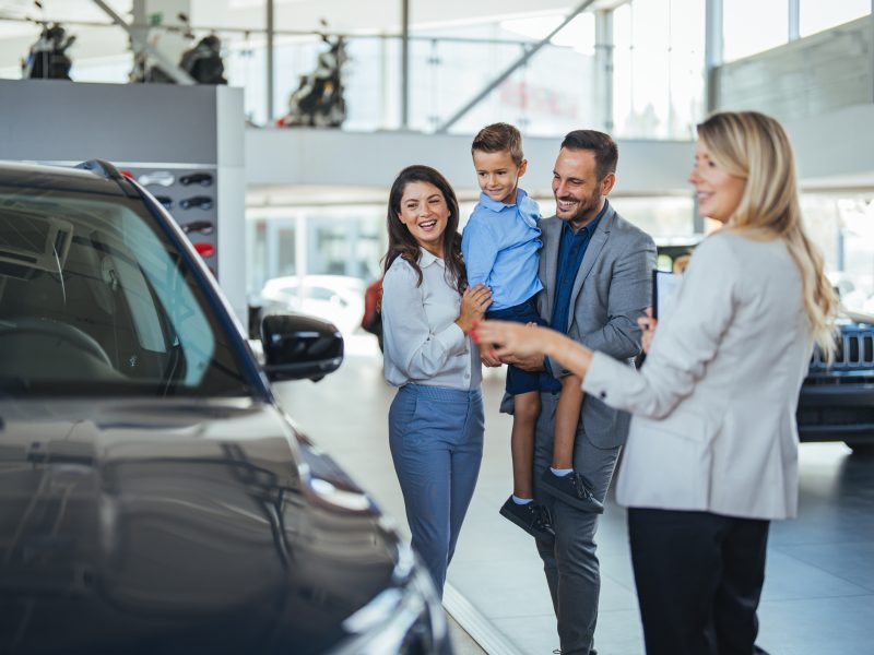 Young happy family enjoying while buying a new car in a showroom. Saleswoman at car dealership center helping family to choose new family vehicle. Family in a car dealership