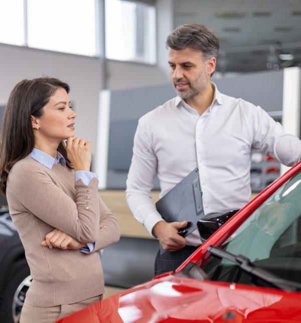 A man and a woman stand beside a red car as they purchase a new vehicle from a salesperson at a car dealership.