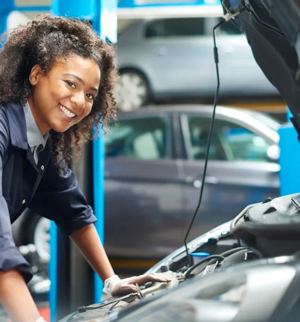 A female mechanic is working under a bonnet of a car in a garage repair shop. She is wearing blue overalls.  She is looking proudly  to camera.