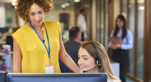 a young call centre representative greets a caller in a large open plan office as her supervisor watches over her shoulder and helps her through the call. Co-workers can be seen defocussed in the background .